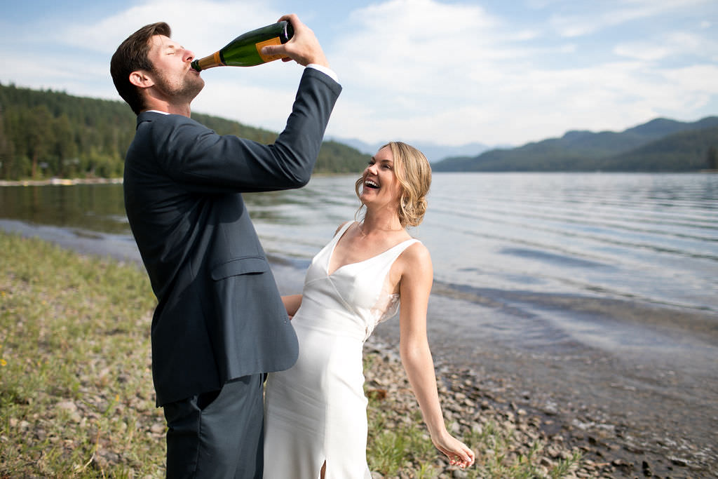 Groom drinking champagne from bottle lakeside while bride smiles