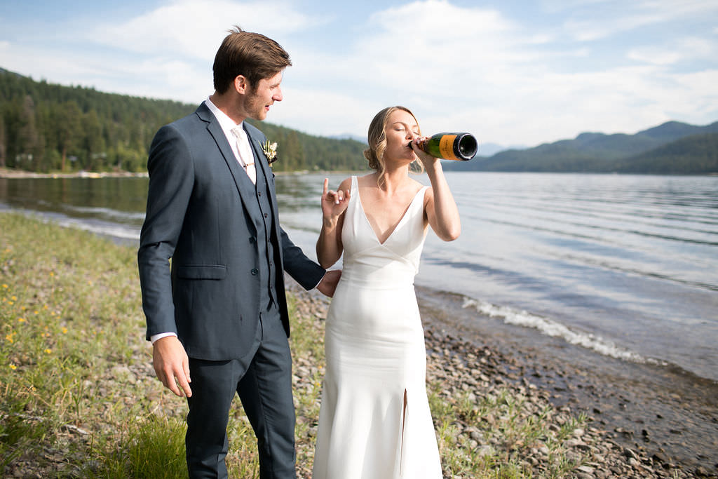 Bride drinking champagne from the bottle lakeside while groom smiles at her 